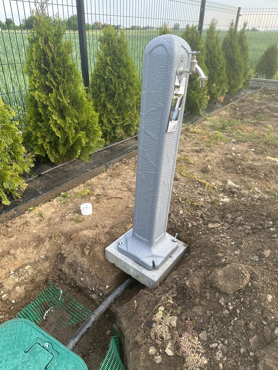 Szara, zewnętrzna kolumna z kranem zamontowana na betonowej podstawie, z widocznymi przyłączami wodnymi w wykopie ziemnym i szeregiem tuj w tle.