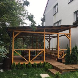 SoBo - Drewniany taras z zadaszeniem, wyposażony w meble ogrodowe, z widocznym zbiornikiem na deszczówkę i młodymi tujami wzdłuż obrzeża.