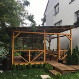 Drewniany taras z zadaszeniem, wyposażony w meble ogrodowe, z widocznym zbiornikiem na deszczówkę i młodymi tujami wzdłuż obrzeża.