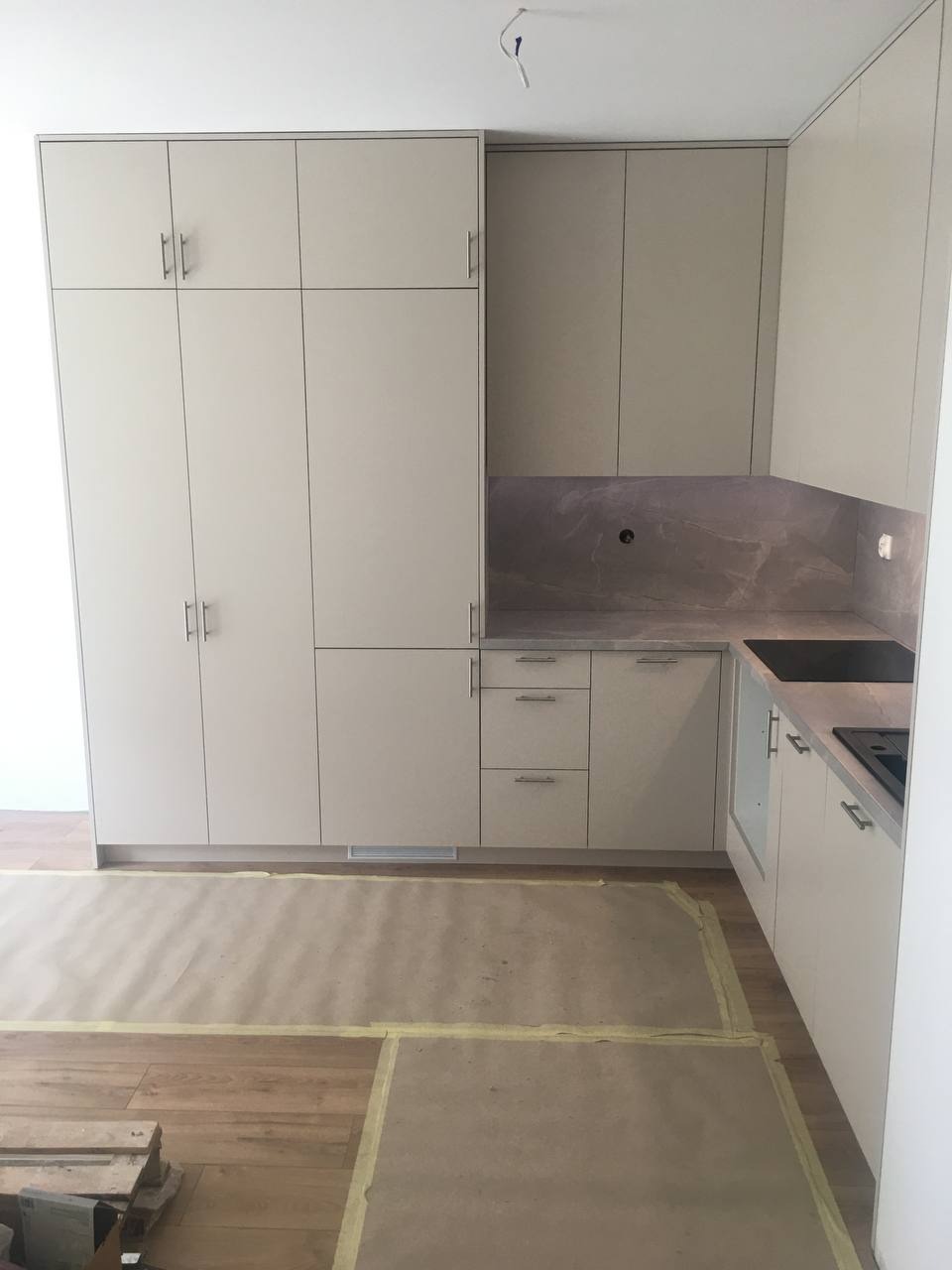 Jasna, nowoczesna zabudowa kuchenna z wysokimi szafkami i marmurowym panelem nad blatem, podłoga zabezpieczona kartonem podczas montażu.