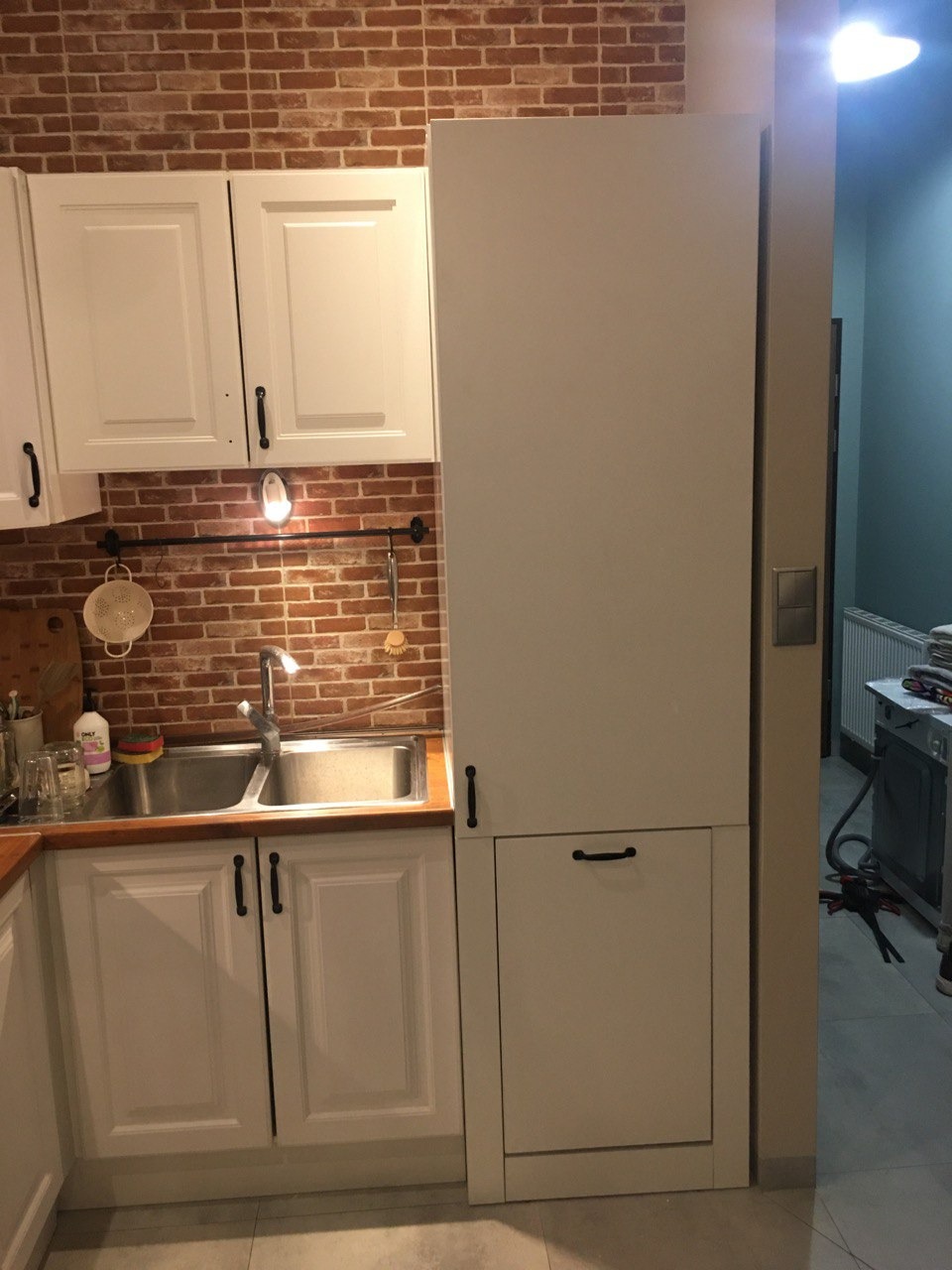 Biała zabudowa kuchenna z ceglaną ścianą w tle, widoczne szafki górne i dolne, zlewozmywak dwukomorowy oraz wysoka szafa zintegrowana z zabudową.