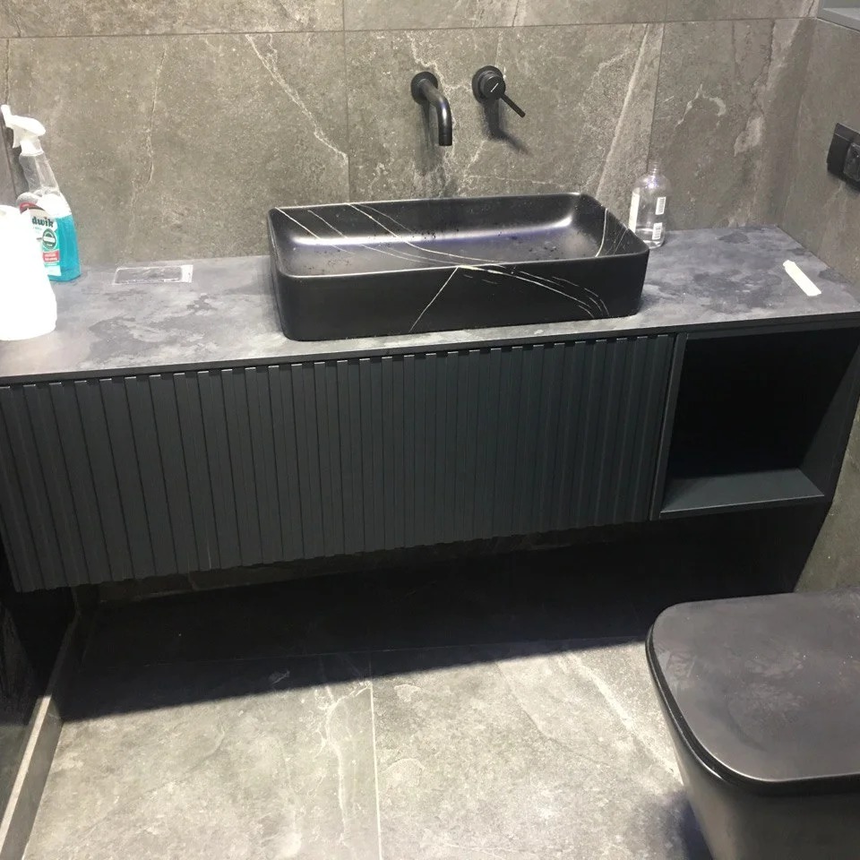 Czarna umywalka nablatowa z marmurowym wzorem, zamontowana na szafce z ryflowanymi frontami w ciemnym kolorze, bateria ścienna, szara marmurowa ściana i podłoga.