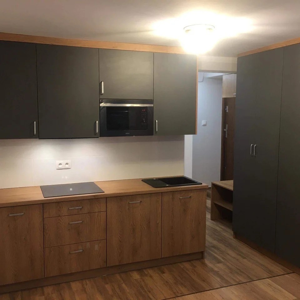 Nowoczesna kuchnia z szarymi szafkami górnymi i drewnopodobnymi szafkami dolnymi, czarnym zlewem i płytą indukcyjną, oświetlona lampą sufitową.