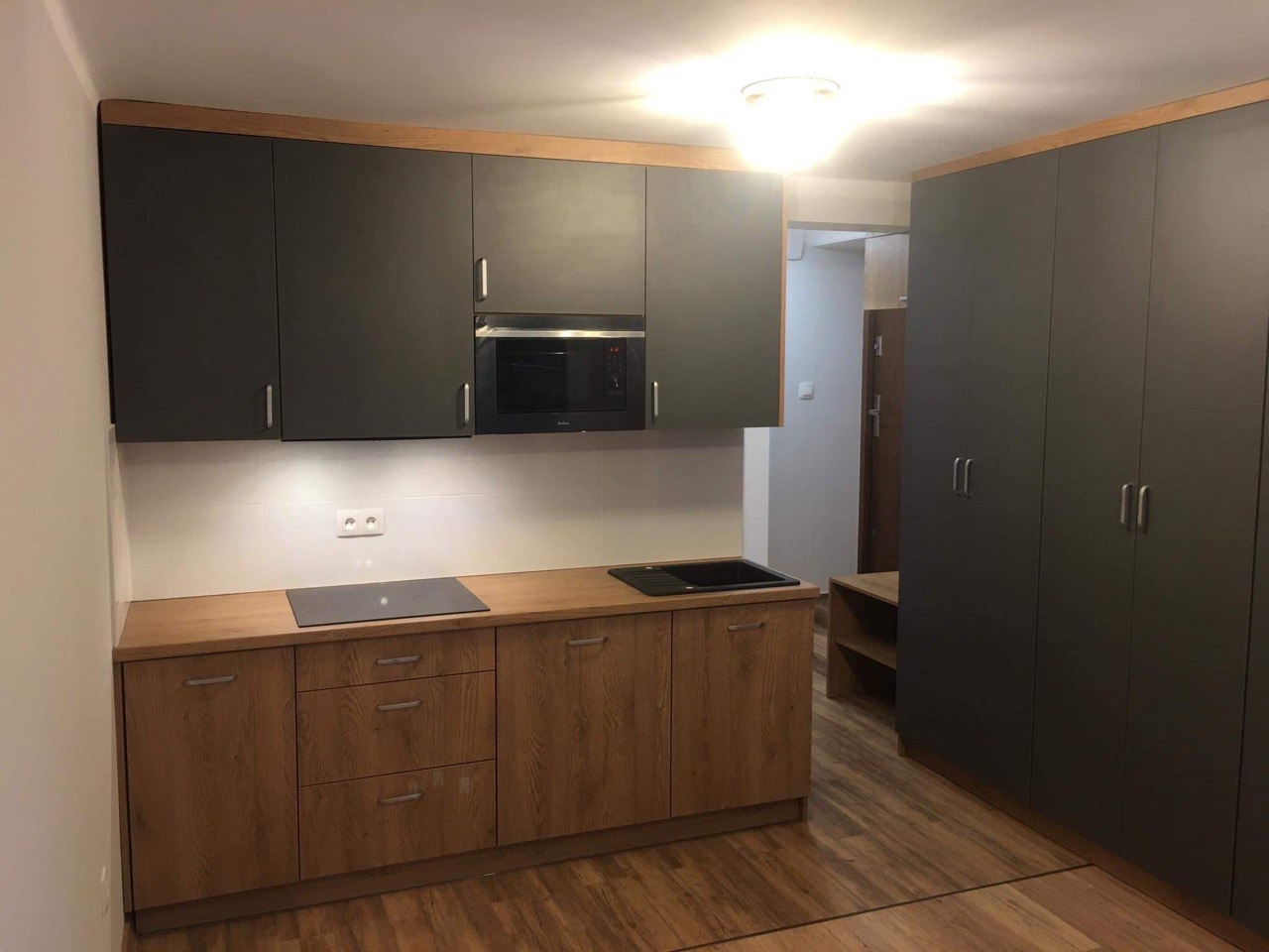 Nowoczesna kuchnia z szarymi szafkami górnymi i drewnianymi szafkami dolnymi, z wbudowaną mikrofalówką i płytą indukcyjną, uzupełniona wysoką zabudową szaf na ścianie bocznej.