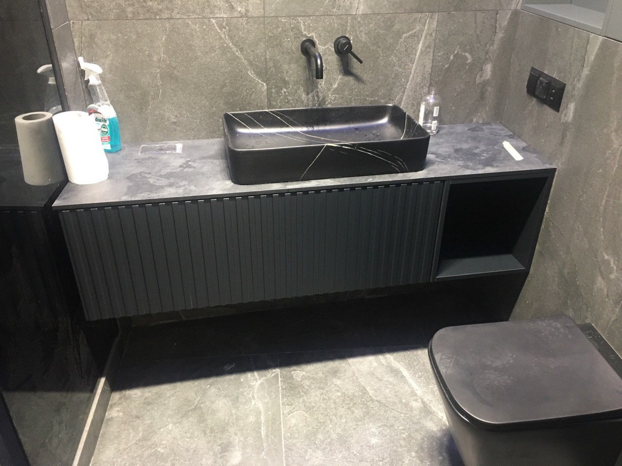 Nowoczesna łazienka z czarną umywalką nablatową o marmurkowym wzorze, szafką pod umywalkę z ryflowanym frontem i szarą płytką imitującą kamień na ścianach i podłodze.