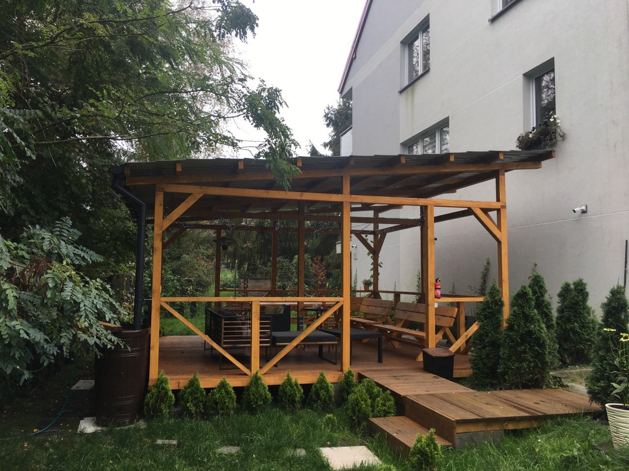 Drewniany taras z zadaszeniem, wyposażony w meble ogrodowe, z widocznym zbiornikiem na deszczówkę i młodymi tujami wzdłuż obrzeża.