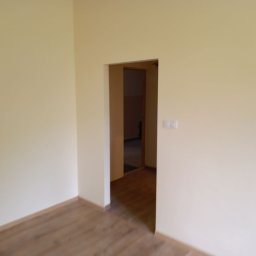 Firma Ogólnobudowlana Andrzej Jackiewicz - Pomalowane na kremowo ściany w pustym pokoju z widoczną podłogą z paneli i otwartym przejściem do kolejnego pomieszczenia.