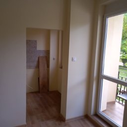 Firma Ogólnobudowlana Andrzej Jackiewicz - Wnętrze pokoju w Suwałkach w trakcie prac przygotowawczych: świeżo pomalowane ściany w odcieniach beżu, drewnopodobna podłoga, widoczny fragment łazienki z częściowo położonymi płytkami i deską...