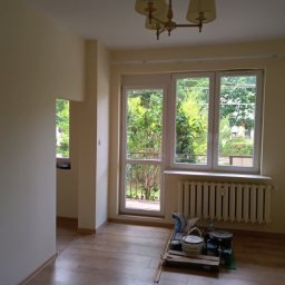 Firma Ogólnobudowlana Andrzej Jackiewicz - Pomalowane na jasnożółto ściany pokoju z oknem balkonowym i grzejnikiem, na podłodze paleta z puszkami farby i akcesoriami malarskimi.