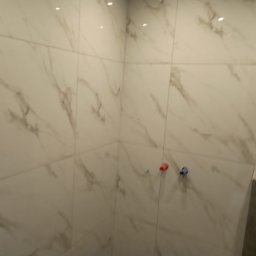 Firma Ogólnobudowlana Andrzej Jackiewicz - Wykończona ściana łazienki z białych, marmurkowych płytek ceramicznych, widoczne wystające rurki doprowadzające wodę z czerwonym i niebieskim oznaczeniem.