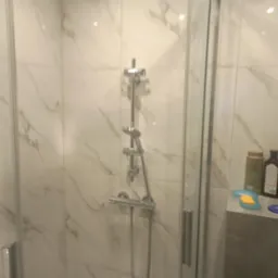 Kabina prysznicowa z chromowaną armaturą i marmurowymi płytkami ściennymi, na półce widoczne kosmetyki i akcesoria łazienkowe.