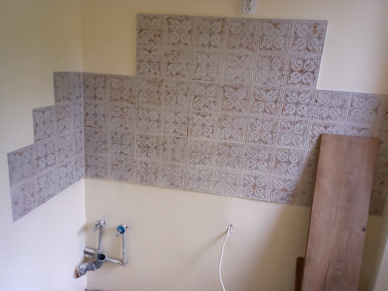 Fragment ściany łazienki z częściowo położonymi płytkami dekoracyjnymi w stylu vintage, widoczne rury hydrauliczne i deska podłogowa oparta o ścianę.