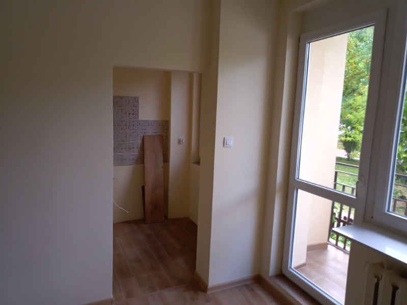 Wnętrze pokoju w Suwałkach w trakcie prac przygotowawczych: świeżo pomalowane ściany w odcieniach beżu, drewnopodobna podłoga, widoczny fragment łazienki z częściowo położonymi płytkami i deską...