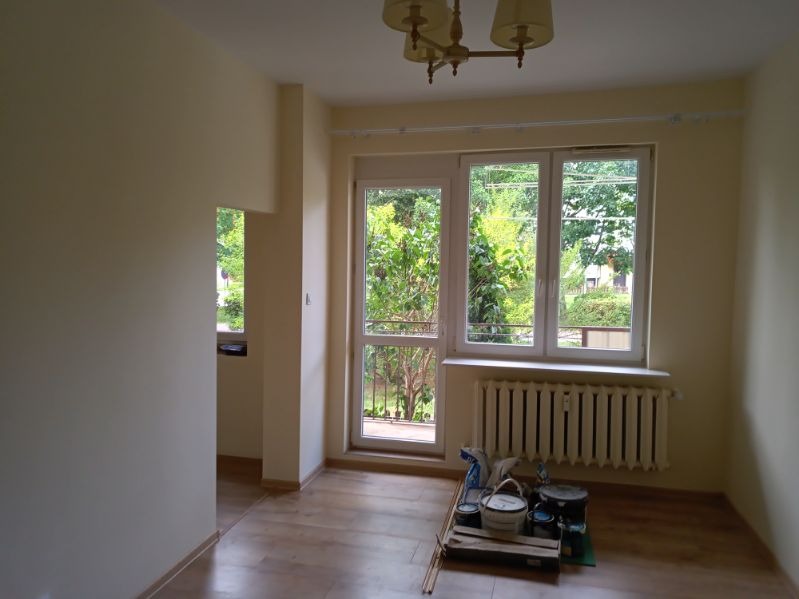 Pomalowane na jasnożółto ściany pokoju z oknem balkonowym i grzejnikiem, na podłodze paleta z puszkami farby i akcesoriami malarskimi.