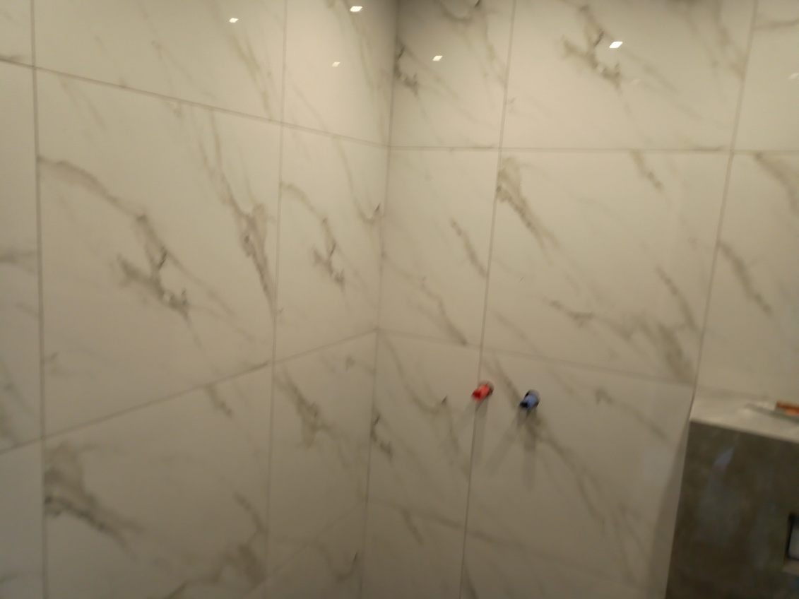 Wykończona ściana łazienki z białych, marmurkowych płytek ceramicznych, widoczne wystające rurki doprowadzające wodę z czerwonym i niebieskim oznaczeniem.