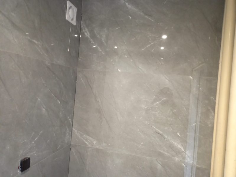 Ściana łazienki wyłożona szarymi płytkami imitującymi marmur, widoczny fragment wentylatora sufitowego z linką, gniazdko elektryczne i fragment rury.