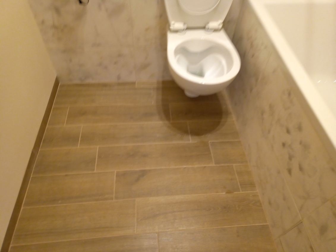 Ukończona łazienka z drewnopodobnymi płytkami podłogowymi i marmurowymi płytkami ściennymi, widoczna toaleta z otwartą klapą i fragment wanny.