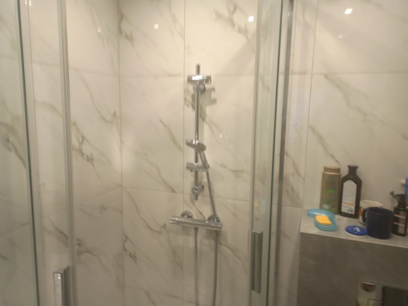 Kabina prysznicowa z chromowaną armaturą i marmurowymi płytkami ściennymi, na półce widoczne kosmetyki i akcesoria łazienkowe.