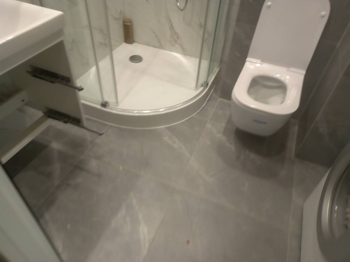 Szara łazienka z kabiną prysznicową z zaokrąglonymi szybami, podwieszaną toaletą i umywalką z otwartą szafką, wykończona płytkami imitującymi marmur.