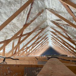 Radpur izolacje natryskowe - Poddasze z drewnianą konstrukcją krokwiową, pokryte grubą warstwą białej pianki PUR, widoczne przewody elektryczne przymocowane do belek.