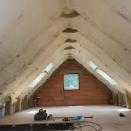 Poddasze z widoczną warstwą izolacyjną z pianki na skośnych ścianach i suficie, małe okno dachowe w ścianie szczytowej z czerwonej cegły, podłoga z płyt OSB, widoczne narzędzia i torba robocza.