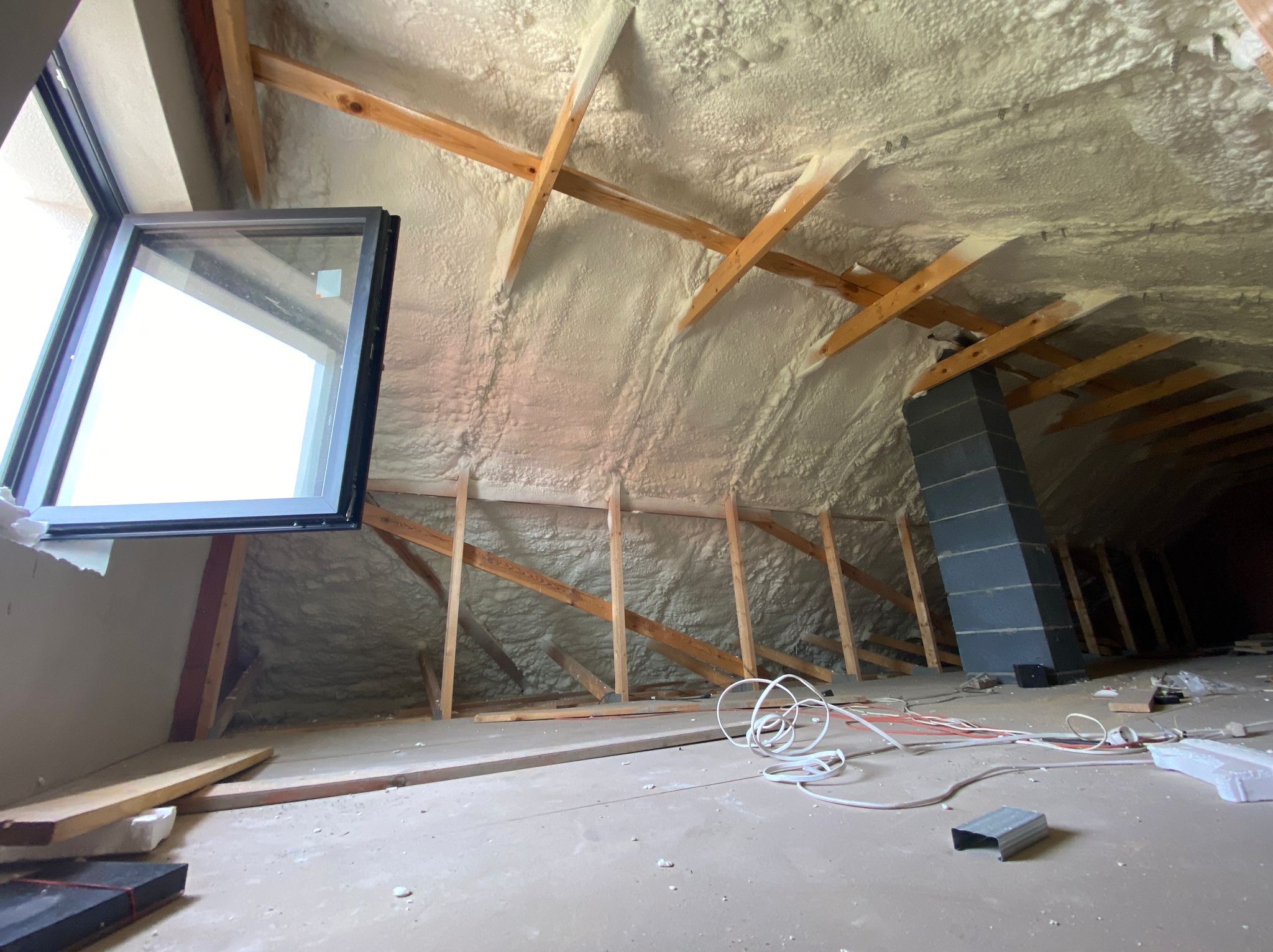 Poddasze po natrysku izolacji pianką, widoczne krokwie i komin, okno dachowe w lewym górnym rogu, przewody elektryczne leżące na podłodze.