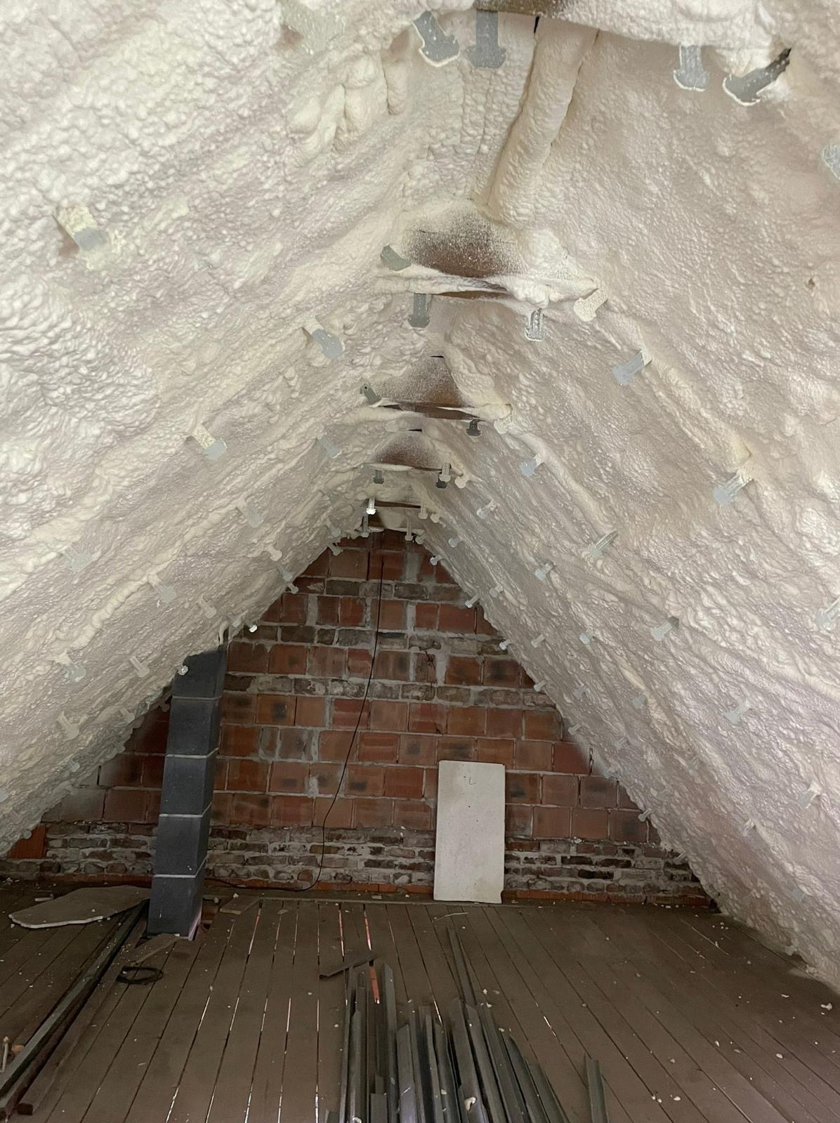 Poddasze z widoczną warstwą izolacyjną z pianki, częściowo zakrywającej krokwie z metalowymi uchwytami, na tle ceglanej ściany szczytowej i drewnianej podłogi z narzędziami.