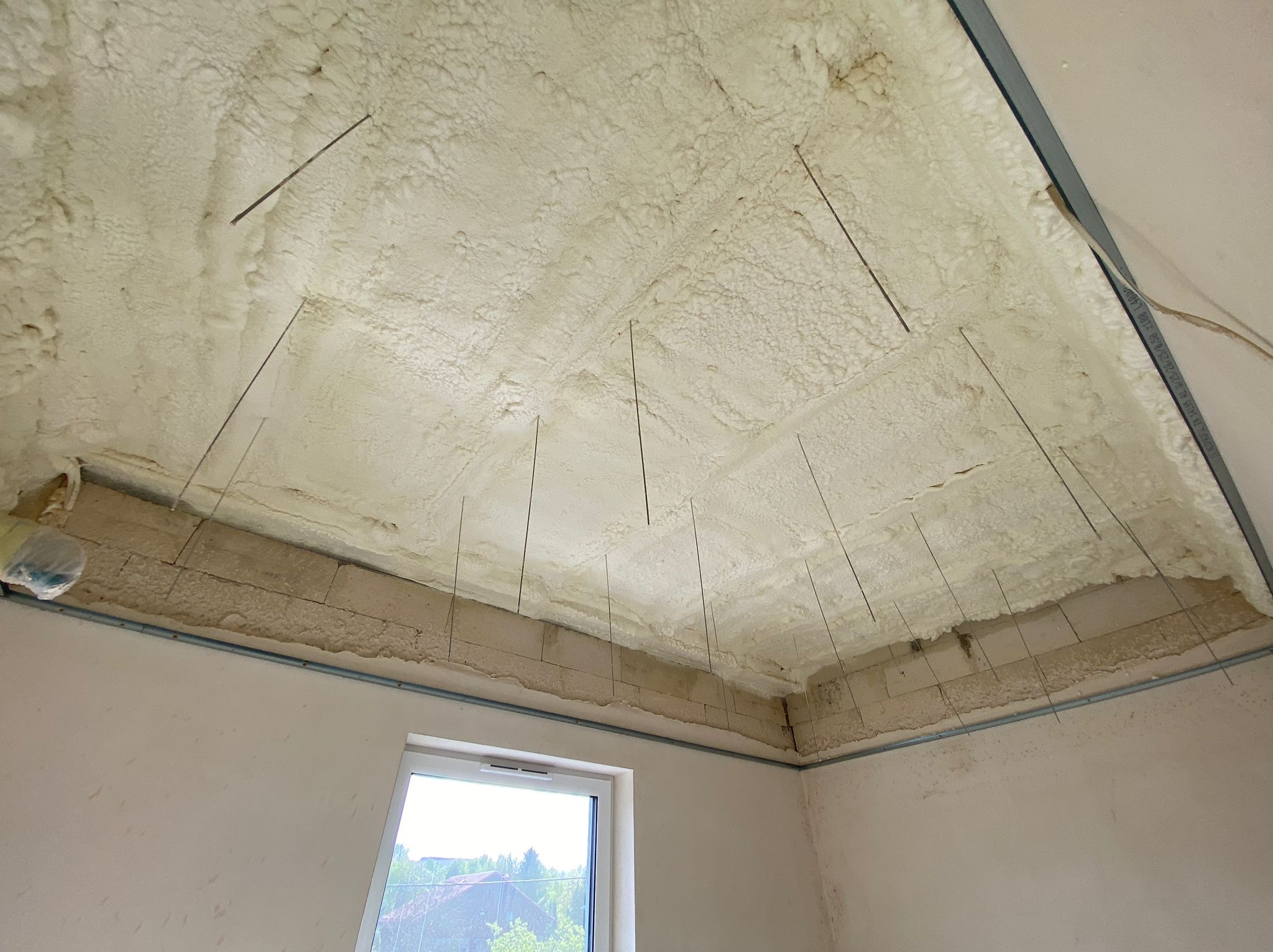 Poddasze po nałożeniu pianki izolacyjnej, widoczne metalowe pręty mocujące, ściany z bloków i fragment okna z widokiem na zewnątrz.