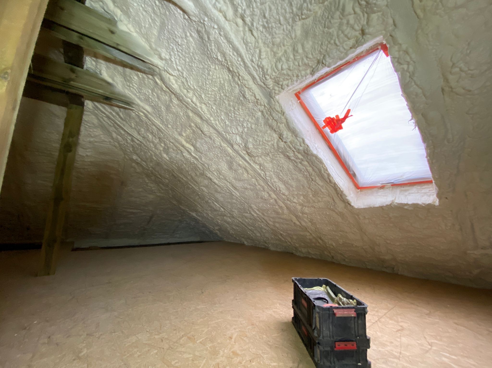 Poddasze po izolacji pianką, widoczne krokwie i okno dachowe zabezpieczone folią, na podłodze skrzynka z narzędziami.