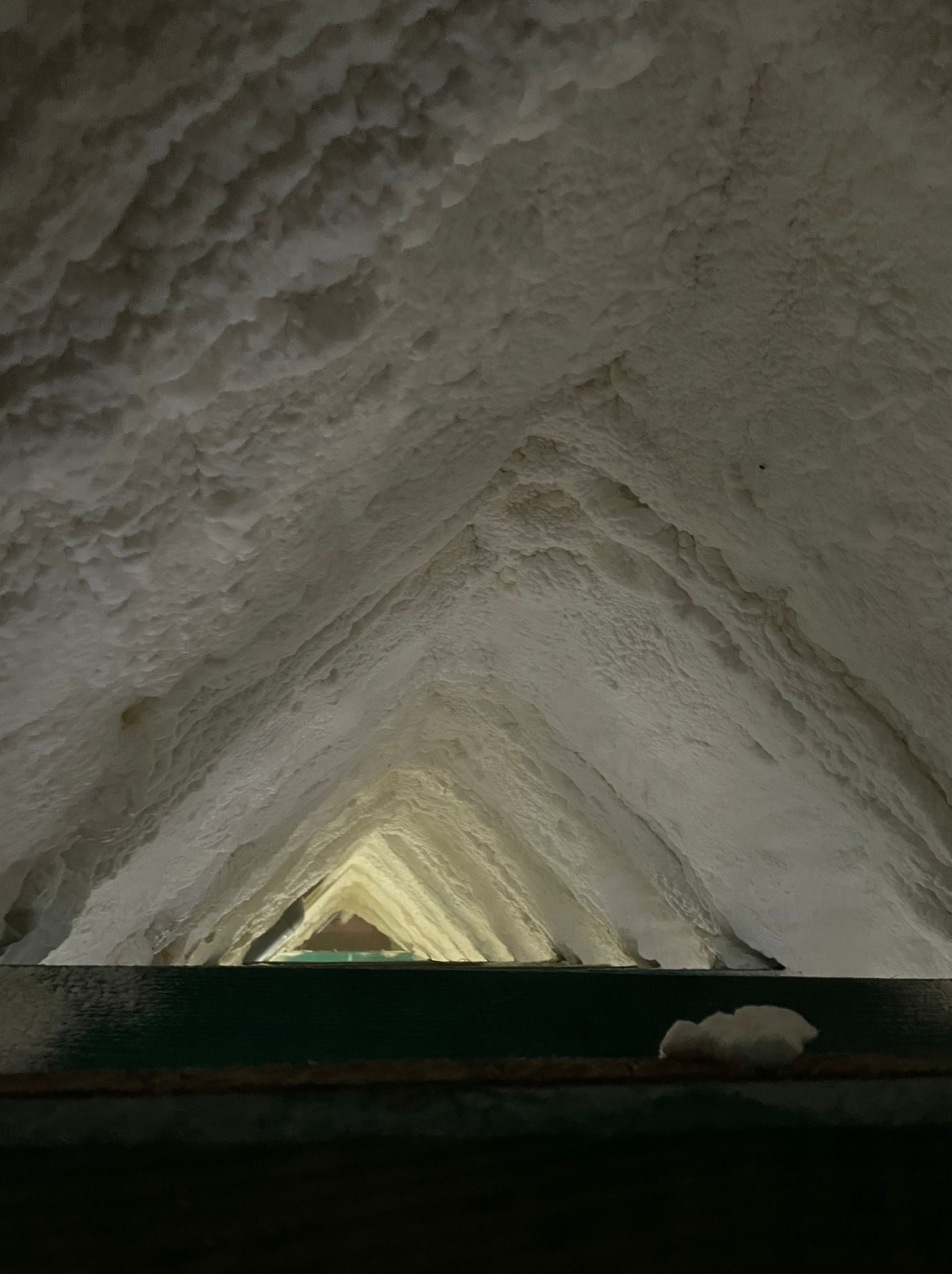 Poddasze ocieplone białą pianką poliuretanową, widok z dołu, ciemne elementy konstrukcyjne w dolnej części kadru, w oddali widoczne szczeliny z dostępem światła zewnętrznego.