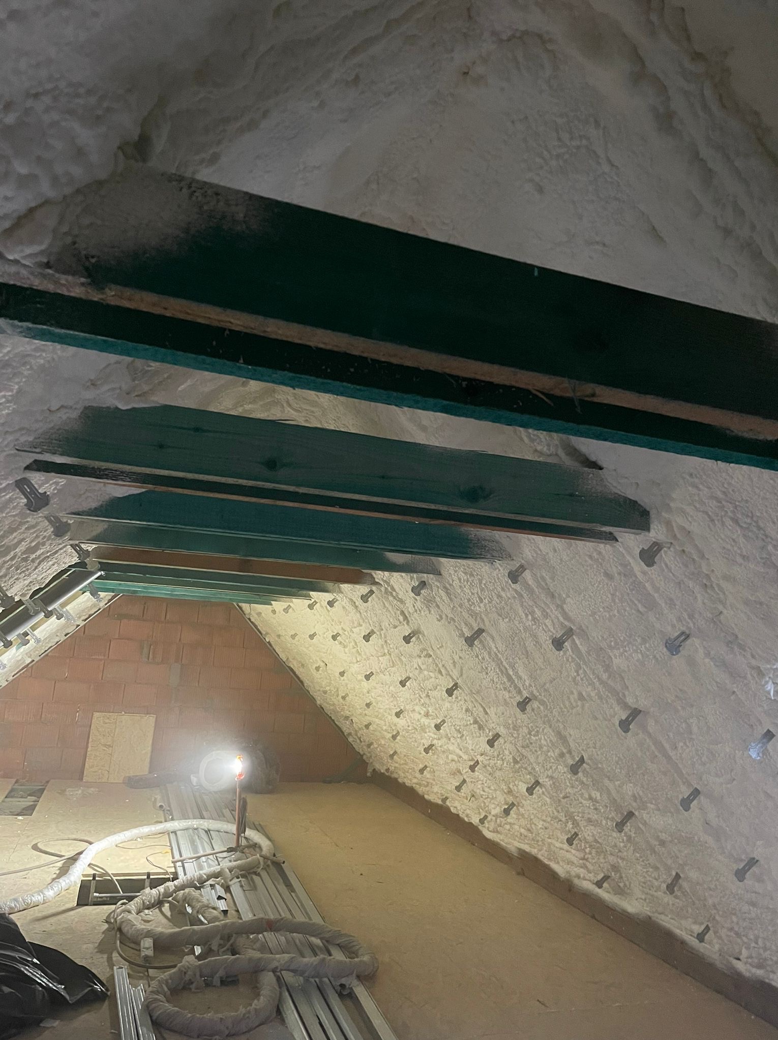 Poddasze z widocznymi krokwiami i izolacją natryskową z pianki, mocowaną na metalowe kołki, oświetlone lampą roboczą.