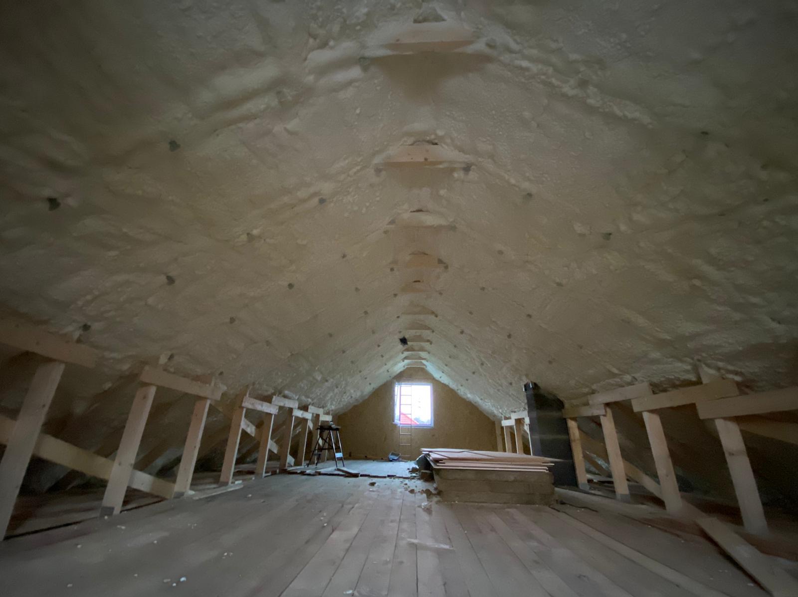 Poddasze z drewnianą konstrukcją pokryte warstwą jasnej pianki izolacyjnej, widoczne okno i drabina.