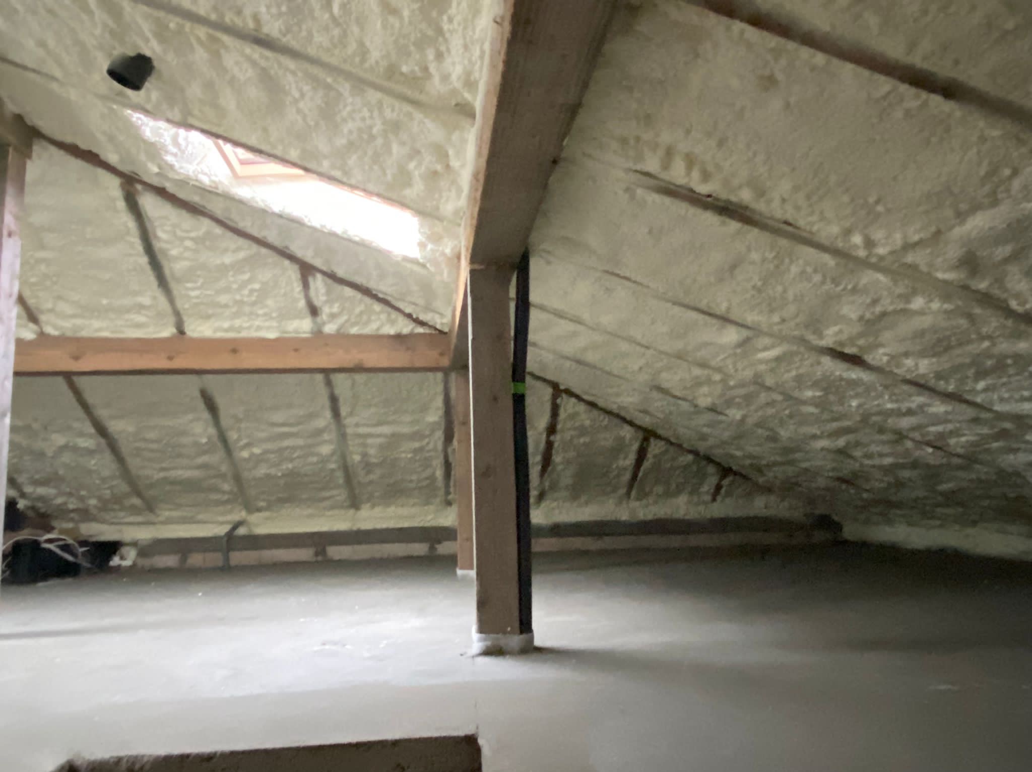 Poddasze po ociepleniu pianką: widoczne drewniane belki konstrukcyjne i biała pianka izolacyjna pokrywająca skośne ściany i sufit, małe okno dachowe w górnej części, czarny przewód przy słupie.
