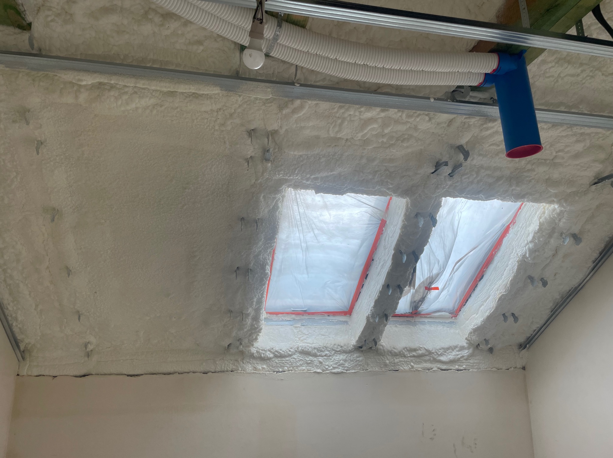 Poddasze po natrysku pianki poliuretanowej, widoczne okna dachowe zabezpieczone folią, elementy konstrukcji metalowej i rury wentylacyjne pod sufitem.