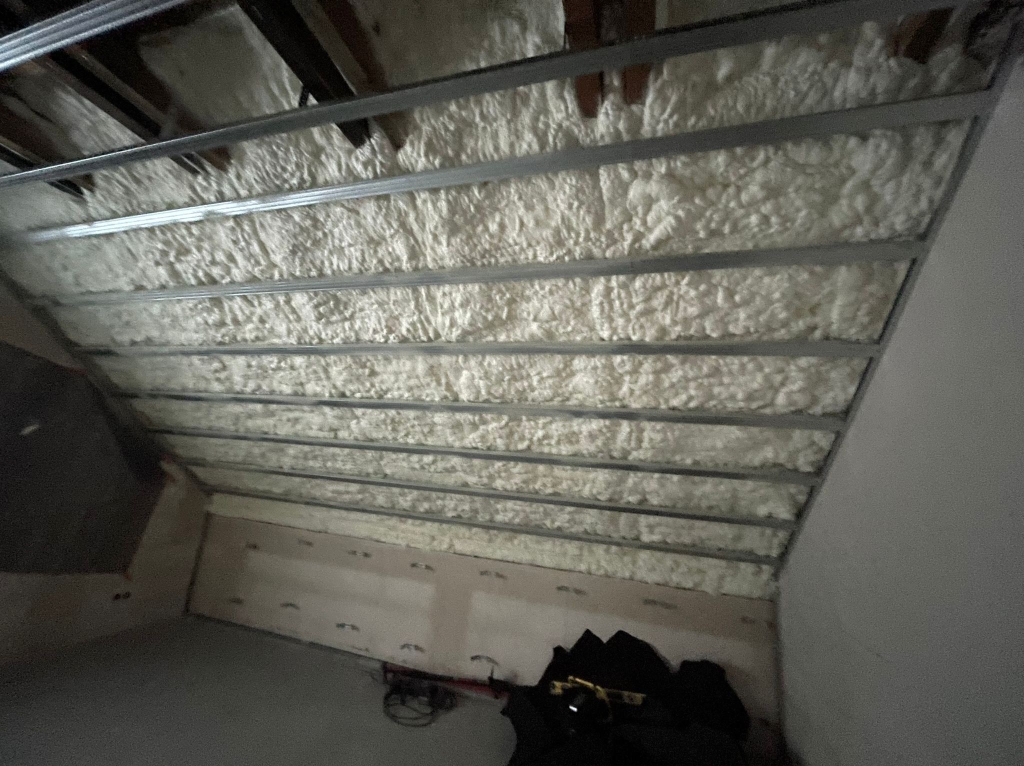 Poddasze ocieplone pianką, widoczne metalowe profile konstrukcyjne, pianka wypełnia przestrzeń między nimi, w tle fragment ściany i podłogi z widocznymi narzędziami.