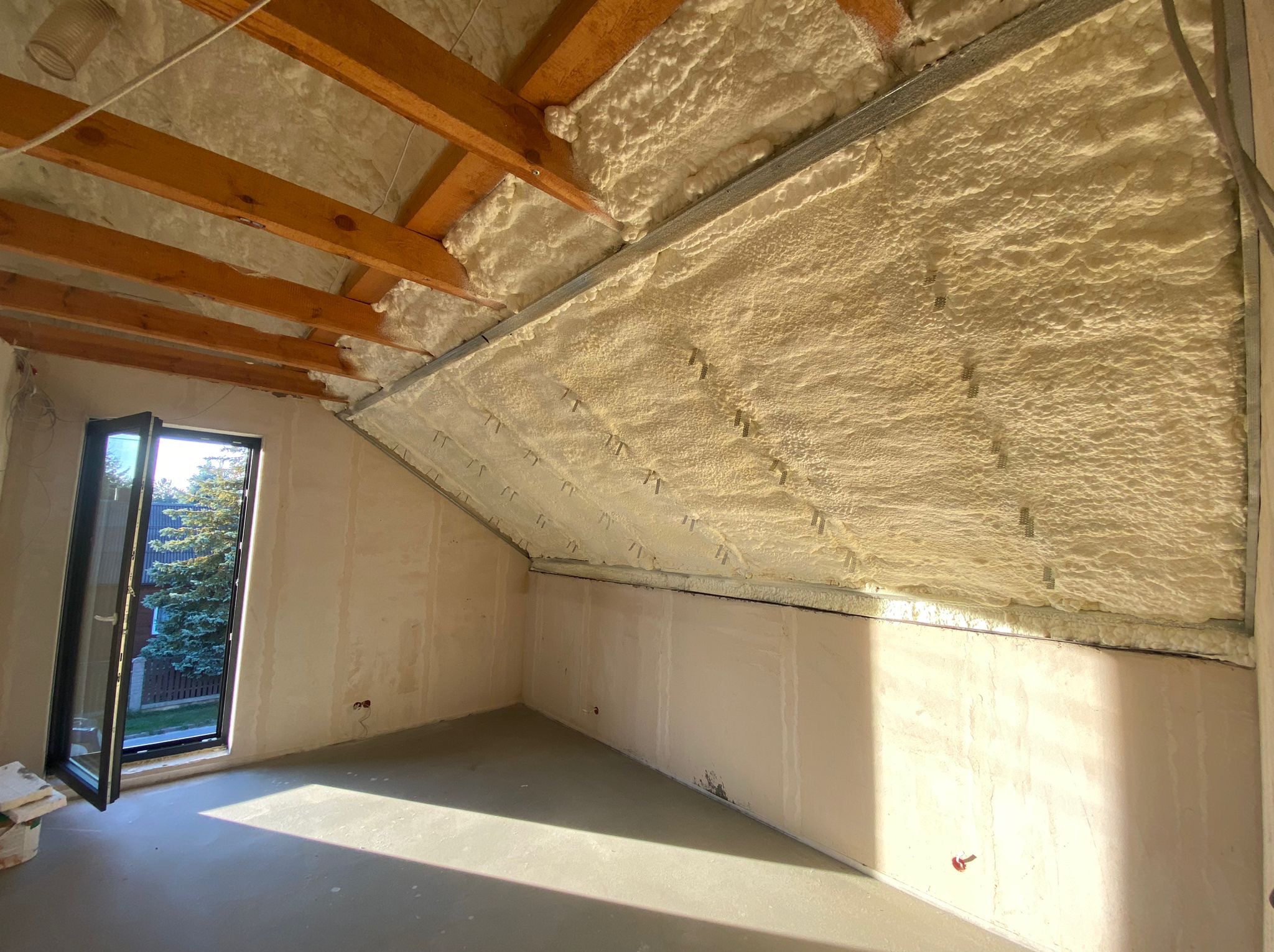 Poddasze po ociepleniu pianką, widoczne drewniane belki stropowe i metalowe profile, ściany przygotowane do dalszych prac wykończeniowych, otwarte okno z widokiem na zieleń.