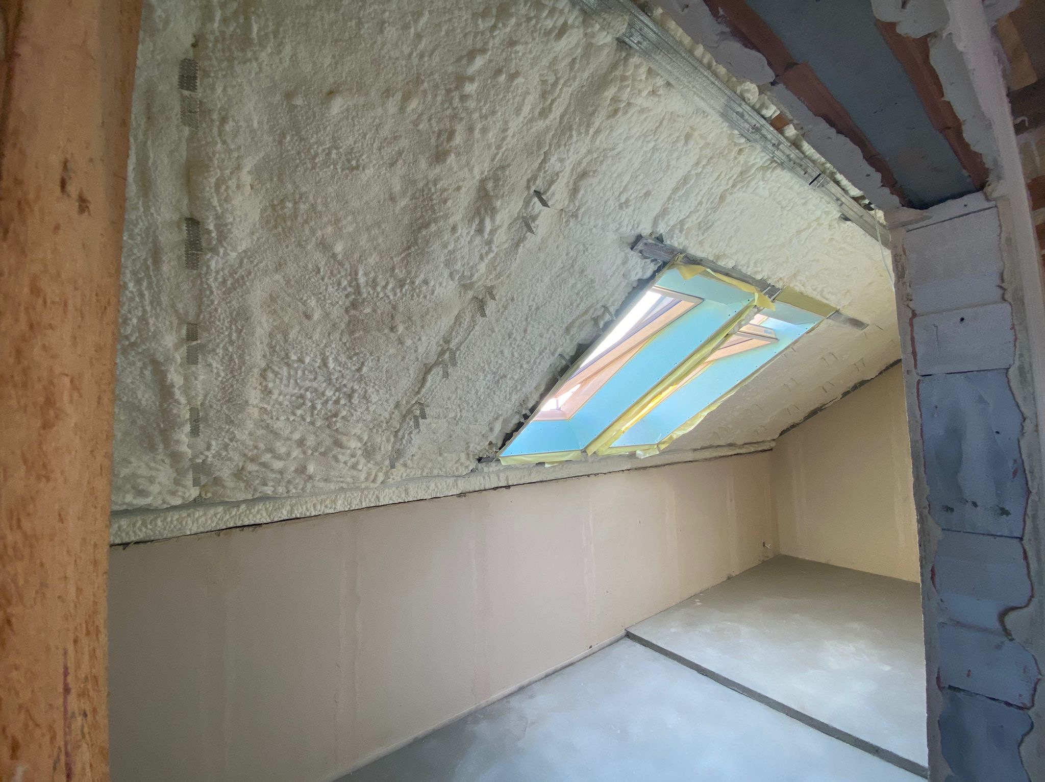 Poddasze po natrysku pianki izolacyjnej, widoczne okno dachowe zabezpieczone taśmą malarską, ślady po montażu profili, oraz fragmenty ścian w trakcie wykańczania.