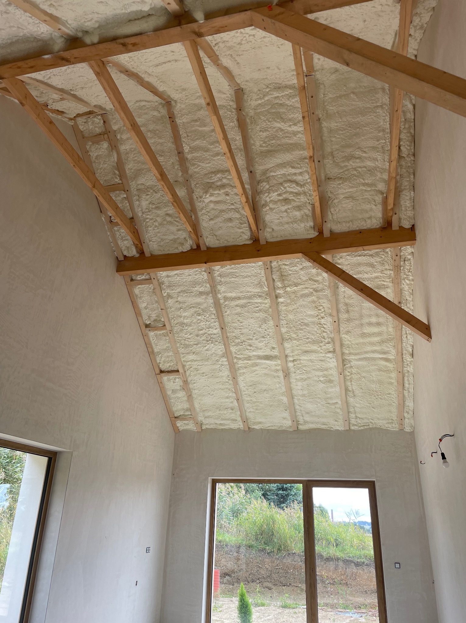 Poddasze domu w trakcie prac izolacyjnych, widoczne drewniane krokwie wypełnione jasną pianką izolacyjną, surowe ściany i okna z widokiem na zewnątrz.