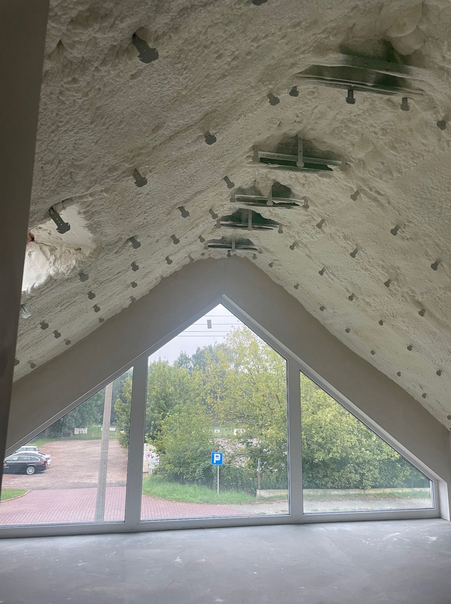 Poddasze po ociepleniu pianką PUR, widoczne metalowe elementy montażowe i trójkątne okno z widokiem na zieleń i parking.