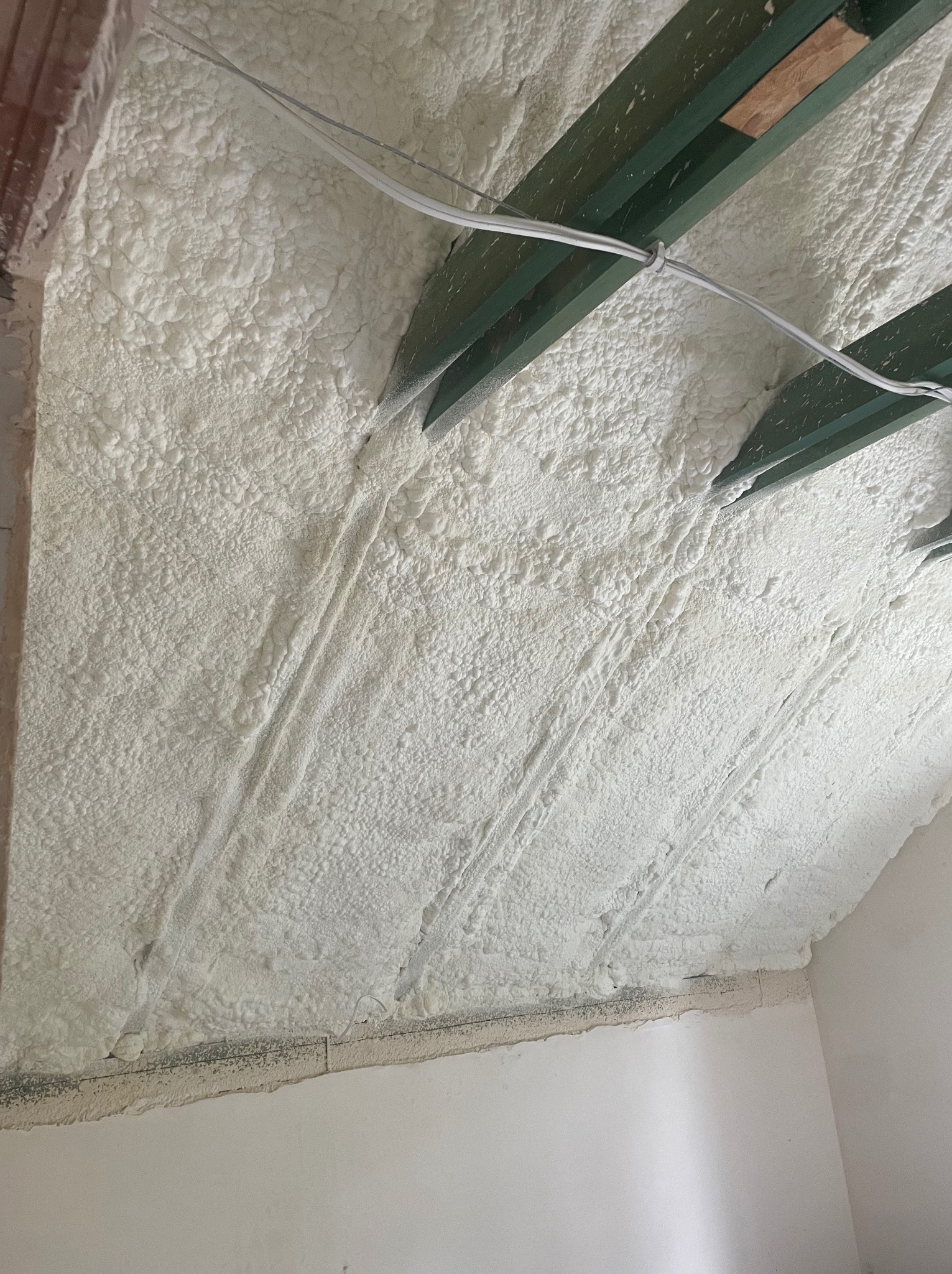 Poddasze z drewnianą konstrukcją pokryte grubą warstwą białej pianki izolacyjnej, widoczne ślady po natrysku i wystające kable elektryczne.