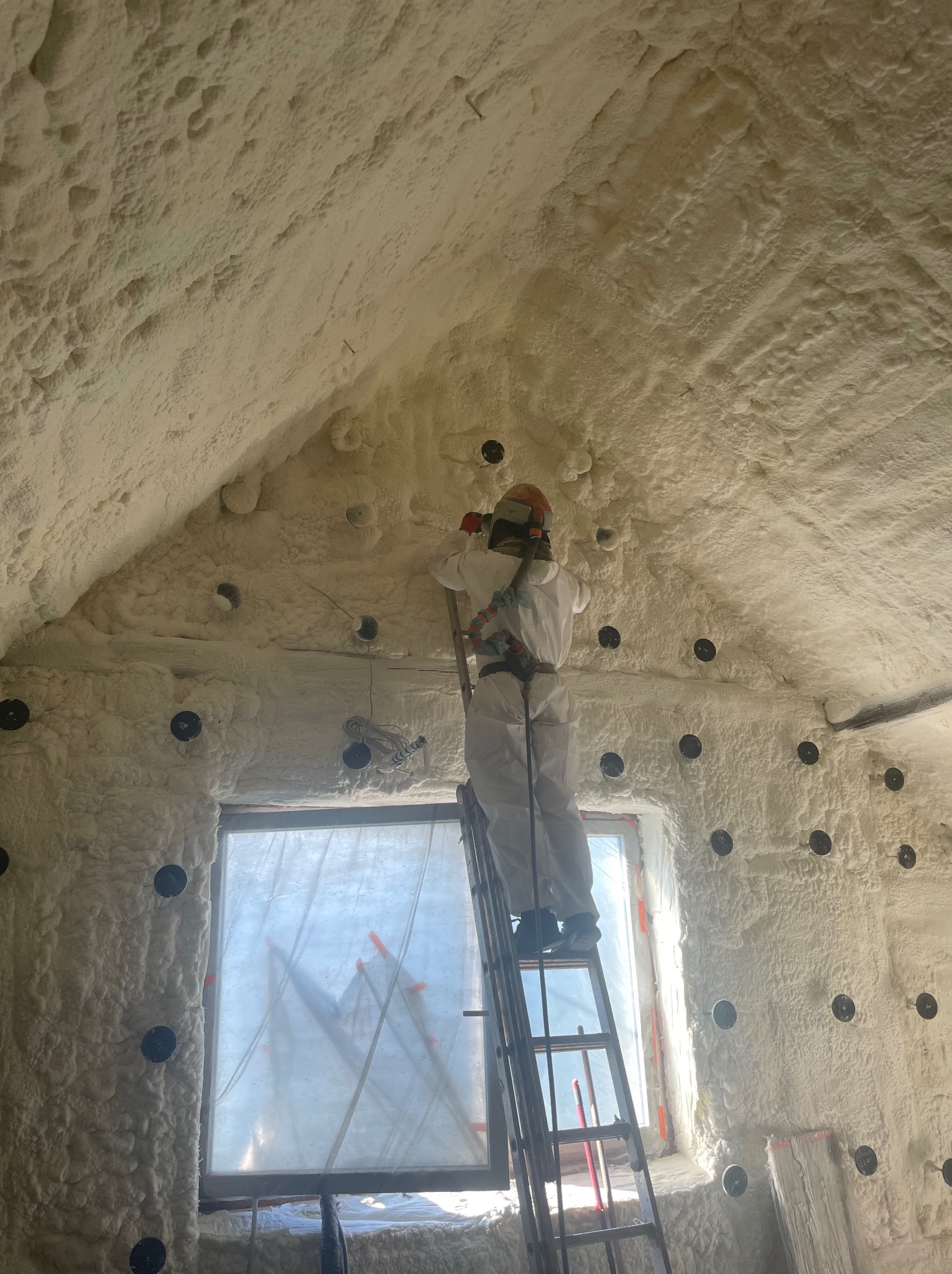Pracownik w kombinezonie ochronnym na drabinie aplikuje piankę izolacyjną na poddaszu w Rudzie Śląskiej, widoczne czarne mocowania i okno zabezpieczone folią.