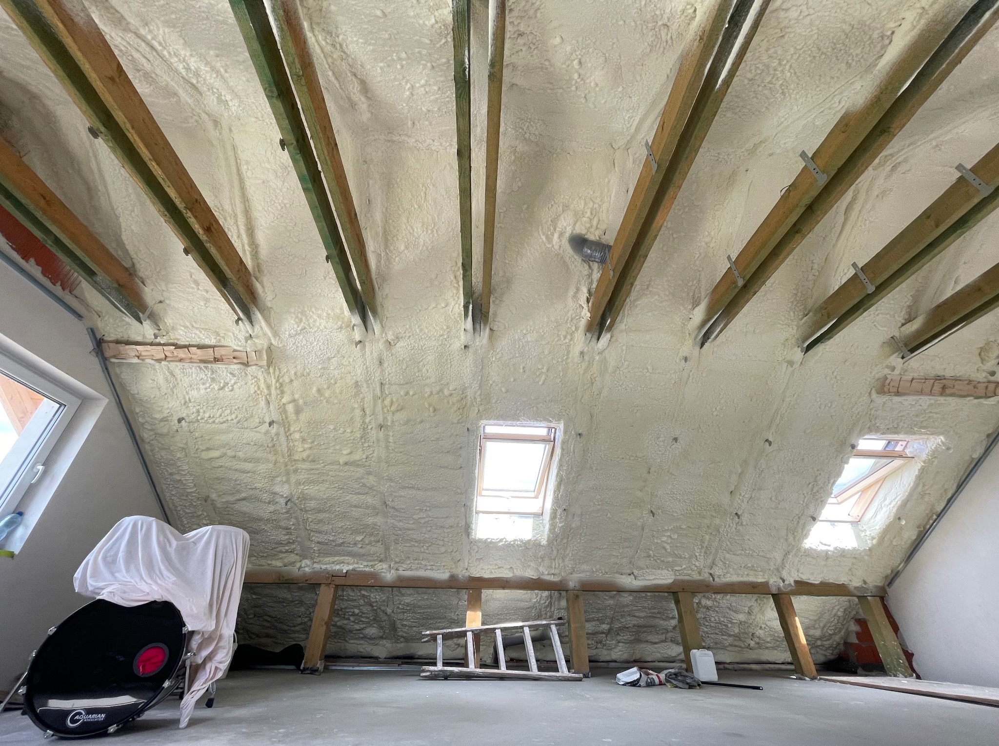 Poddasze po ociepleniu, widoczne drewniane krokwie i okna dachowe, na podłodze drabina i przykryty materiałem zestaw perkusyjny.