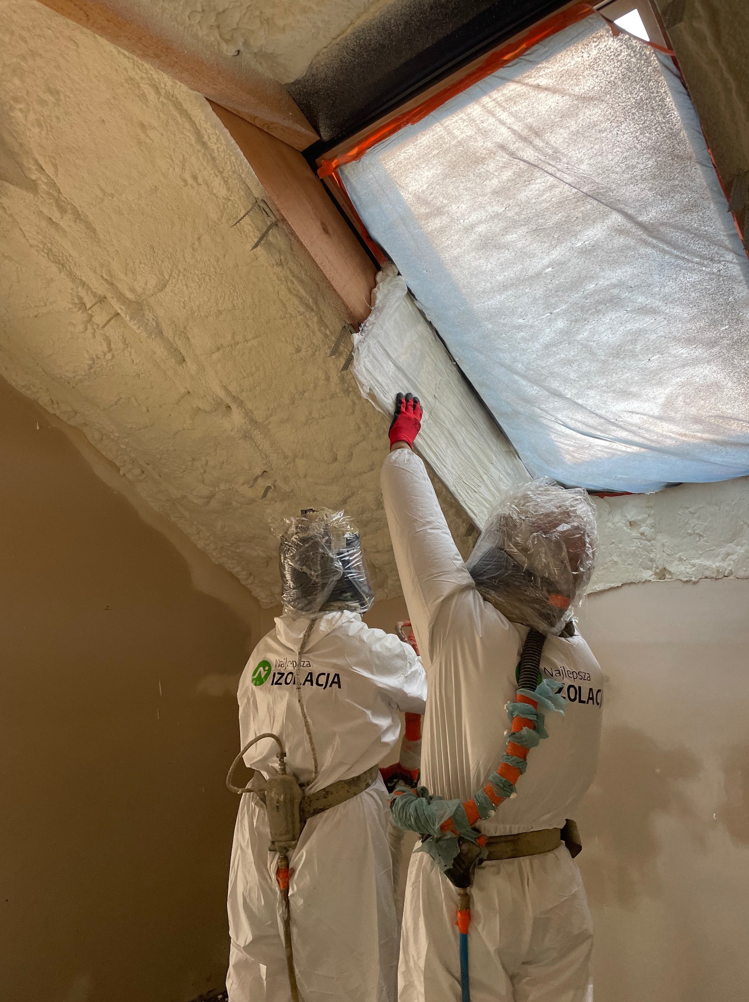 Pracownicy w kombinezonach ochronnych aplikują piankę izolacyjną na poddaszu, widoczne świeżo nałożona pianka i okno dachowe z folią ochronną.