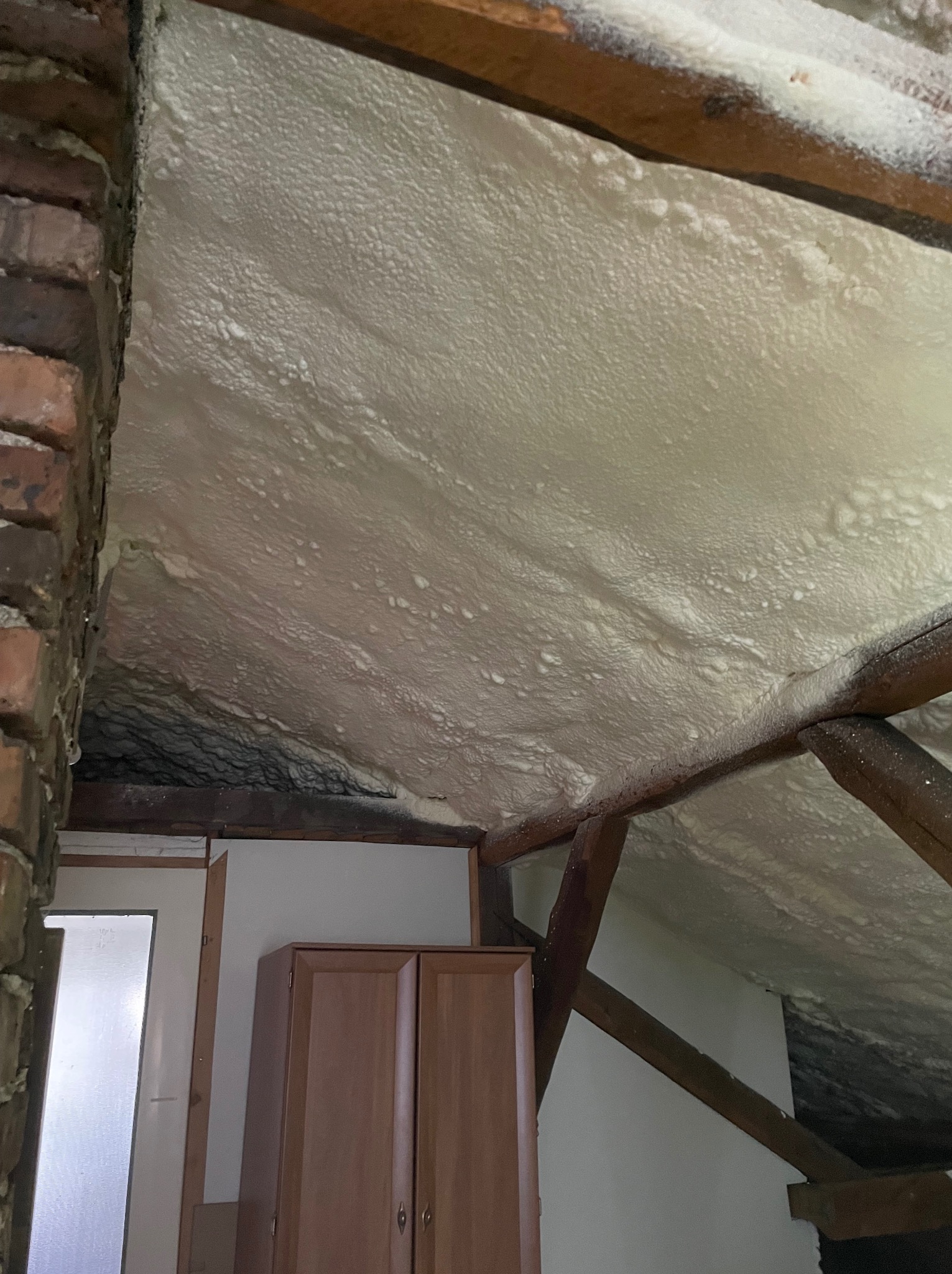 Poddasze z widocznymi krokwiami i ociepleniem natryskowym, fragment ściany z cegły i szafki w tle.