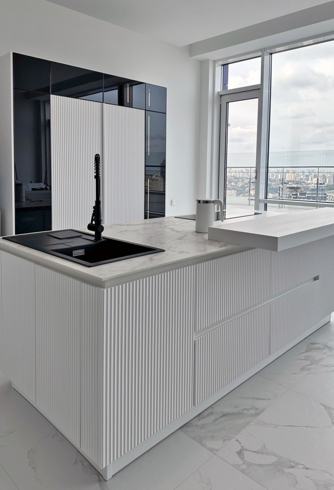 Nowoczesna, biała wyspa kuchenna z ryflowanymi frontami i czarnym zlewem, marmurowy blat, widok na miasto z okna.