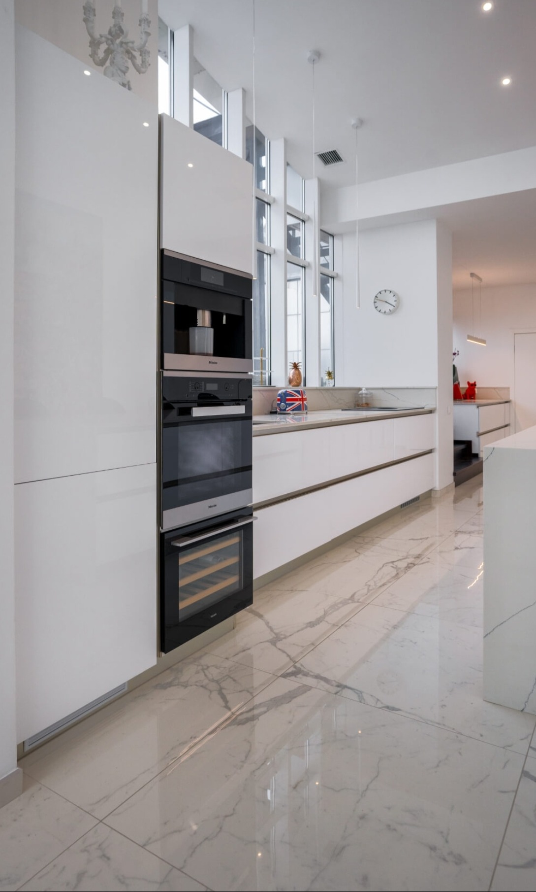 Nowoczesna kuchnia z białymi, lakierowanymi frontami, zabudowanym ekspresem do kawy Miele, piekarnikiem i chłodziarką do wina, marmurowa podłoga odbijająca światło z okien.