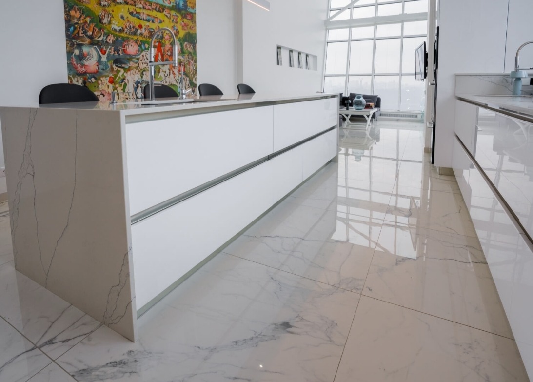 Nowoczesna, minimalistyczna kuchnia z białymi szafkami i blatem imitującym marmur, odbijająca się w lśniącej podłodze, z widocznym fragmentem salonu i dużym oknem w tle.