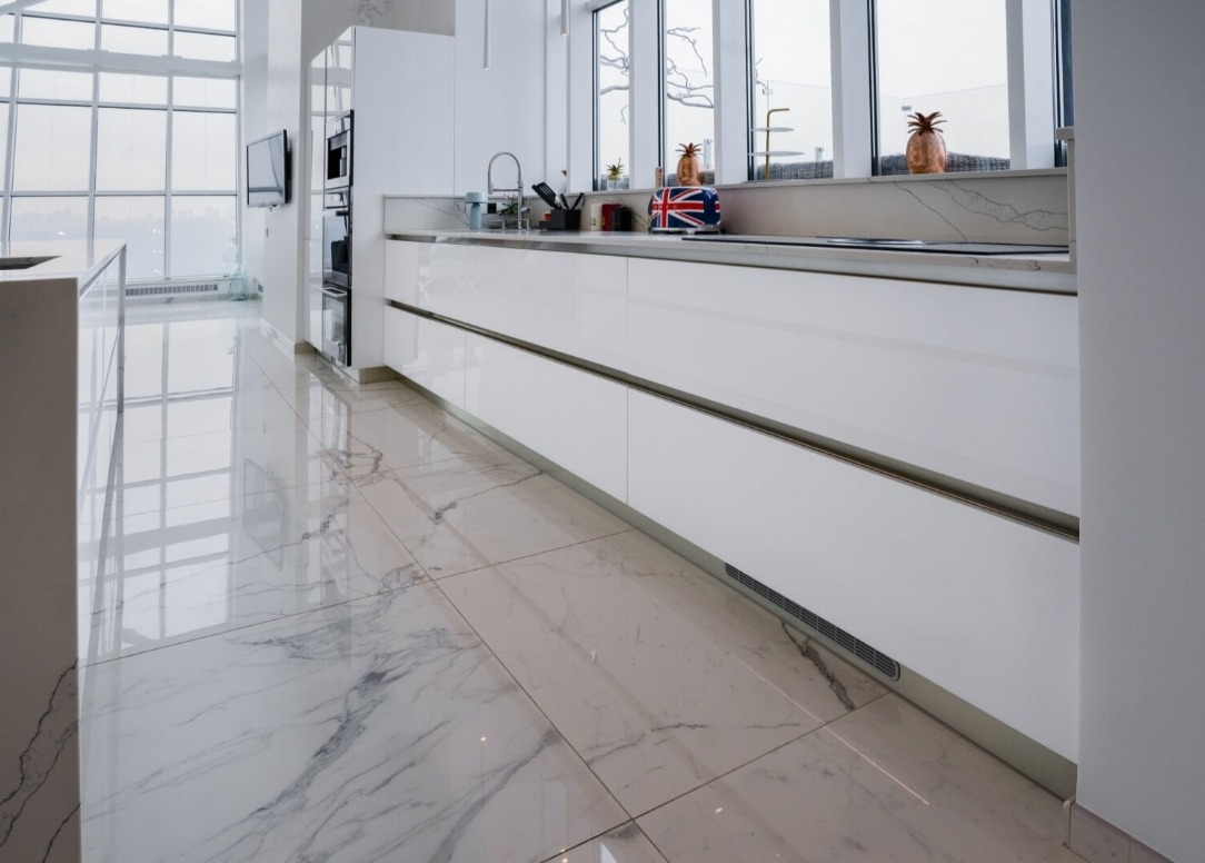 Nowoczesna, biała kuchnia z błyszczącymi frontami szafek i marmurową podłogą, widok na okno z dekoracjami w postaci ananasów i tostera z flagą Wielkiej Brytanii.