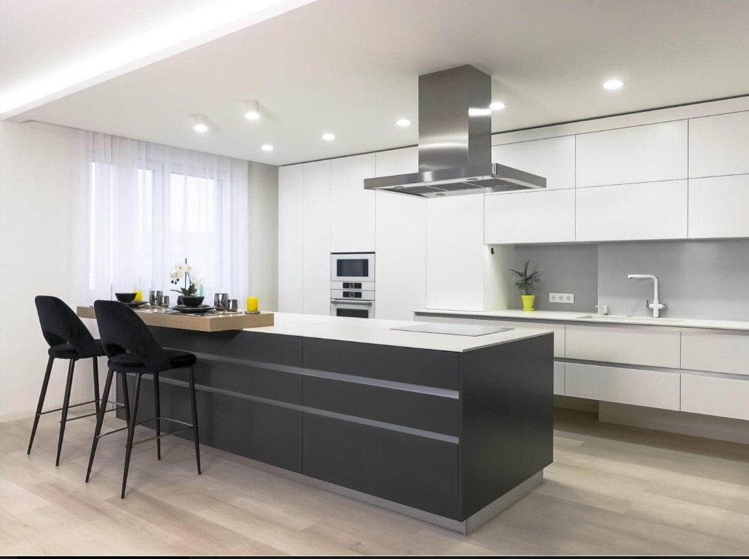 Nowoczesna kuchnia z wyspą w odcieniach bieli i szarości, z dekoracyjnym okapem nad płytą grzewczą i czarnymi hokerami przy blacie.