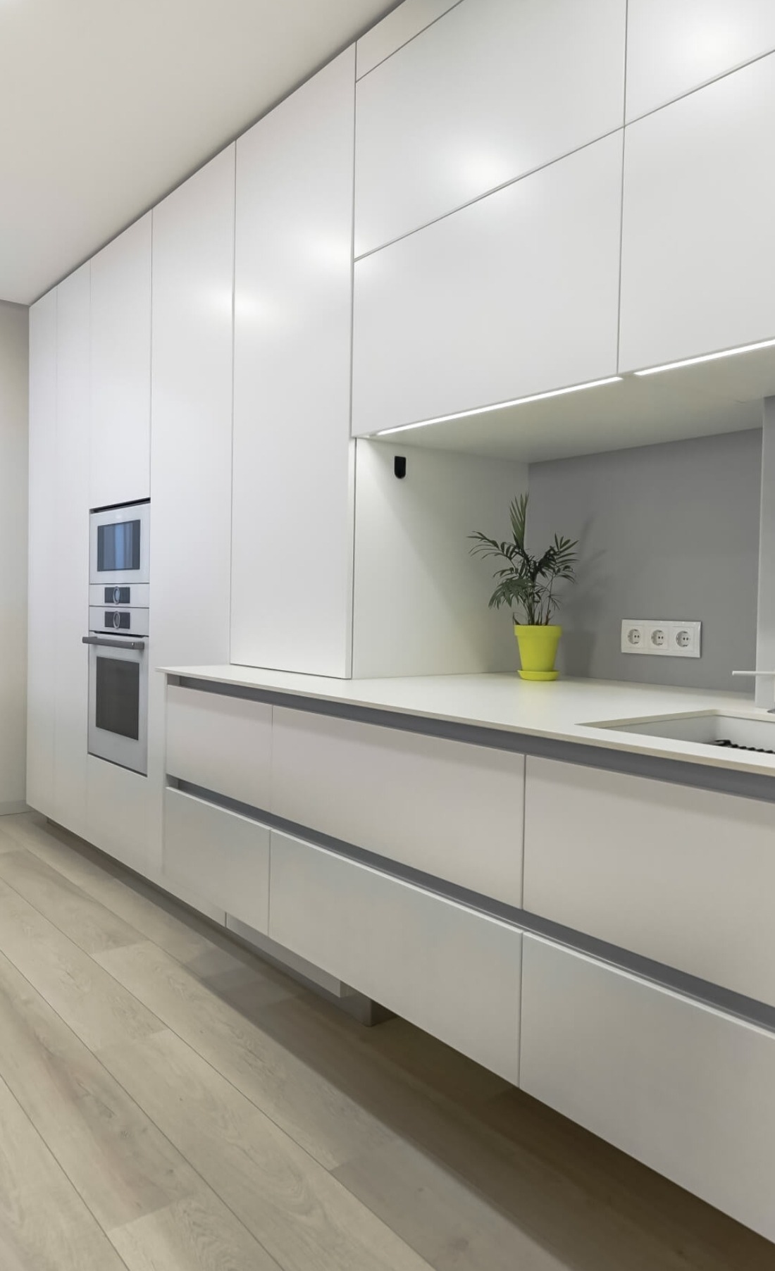 Nowoczesna, minimalistyczna kuchnia z białymi, gładkimi frontami szafek, wbudowanymi piekarnikami i zieloną rośliną w doniczce na blacie.